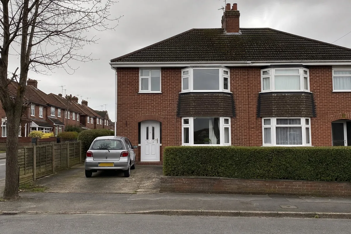 Overcast phone photo of a brick semi-detached house taken square-on from the pavement: a short drive with a silver hatchback, a white front door, two bay windows, a low hedge, and a bare tree at the left edge