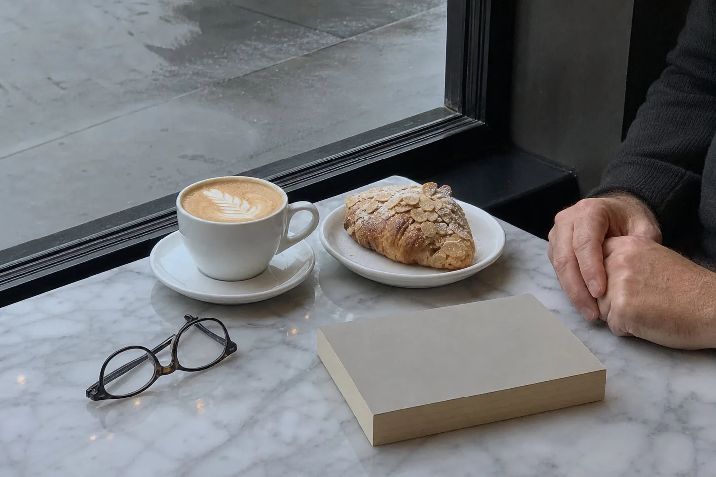 Phone photo of a marble café table by a window with a flat white, an almond croissant, reading glasses, a paperback, and a man's hands, in grey window light