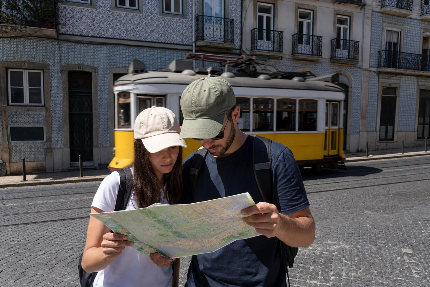 Midday phone photo of a couple in caps reading a map on a cobbled old-town street as a yellow tram passes behind them, with hard shadows and plain digital colour