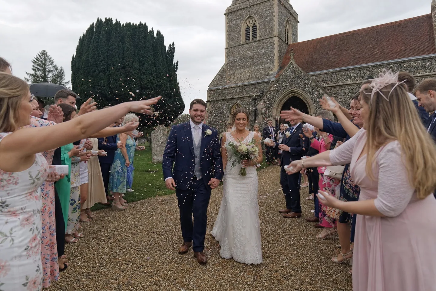 Phone photo of a bride and groom walking down a gravel church path under an overcast sky as guests throw confetti, in flat grey light with washed-out colour