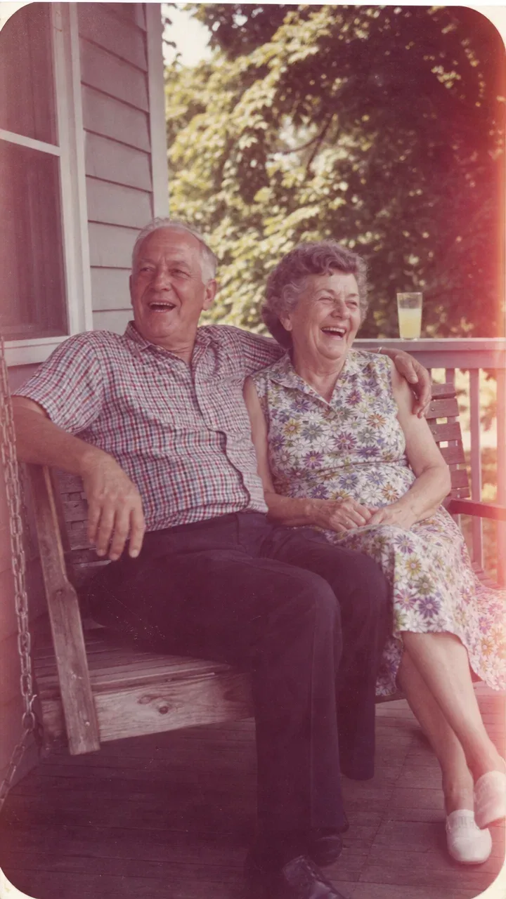 Faded 1970s snapshot of an elderly couple laughing on a wooden porch swing