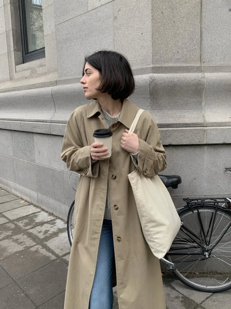 Candid phone photo of a young woman with a short dark bob in a beige trench coat and jeans on a city pavement, holding a plain canvas tote bag and a coffee cup, with a grey stone building and a parked bicycle behind her