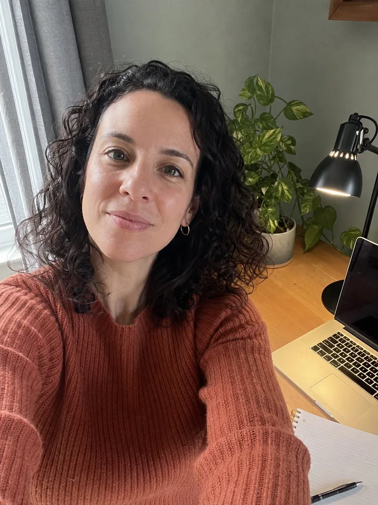 Arm’s-length front-camera selfie of a woman with curly dark hair and hoop earrings in a rust ribbed jumper at a home desk, a laptop, notebook and pothos plant behind her, window light and a desk lamp