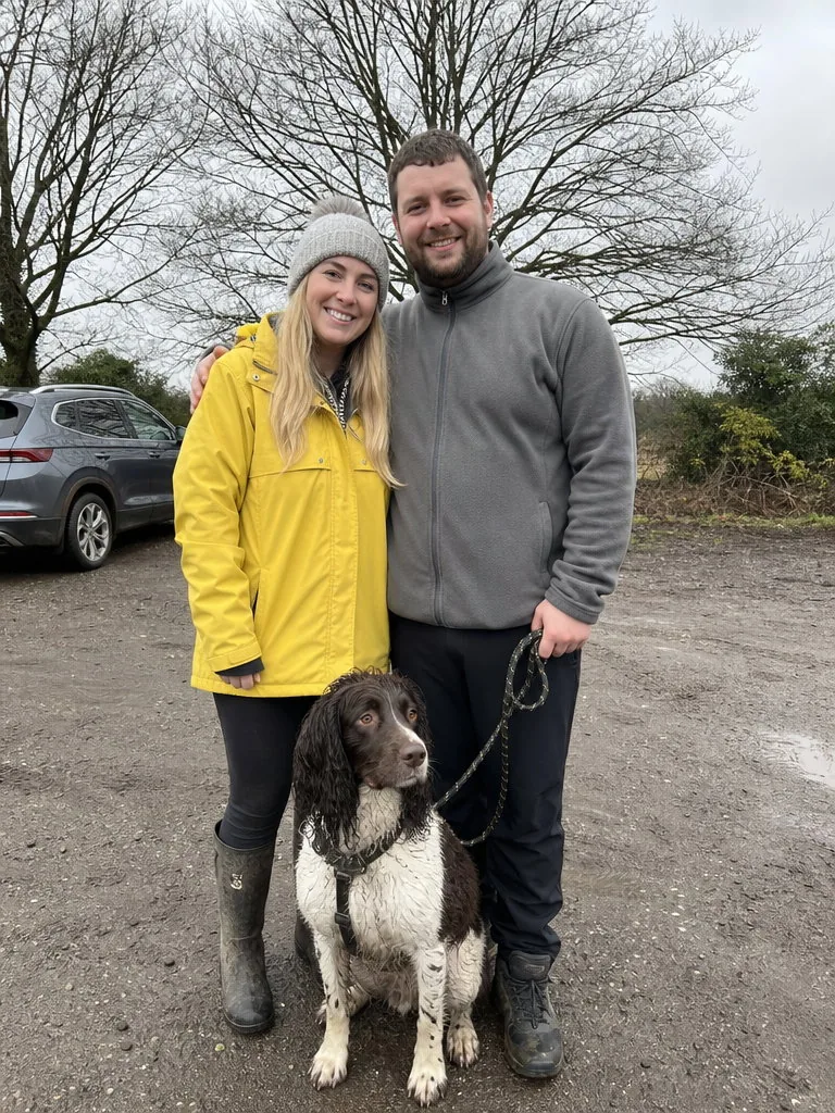 Flat grey-day phone photo of a couple in a yellow raincoat and a grey fleece holding a wet dog on a lead in a muddy trailhead car park, a parked car and bare trees behind