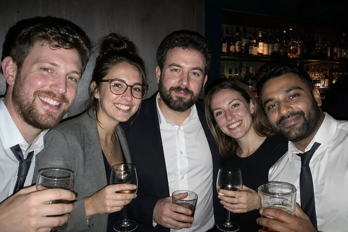 Flash snapshot of five coworkers crowded together holding drinks at a leaving-do in a dim bar, hard flash shadows on the wall and bottles on the shelves behind