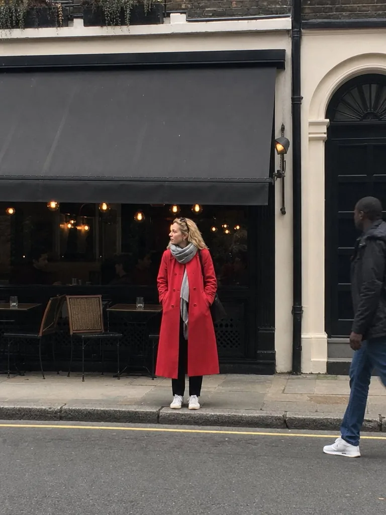 Phone photo taken from across the street of a woman in a red wool coat and white sneakers standing small outside a cafe with a dark awning, a passer-by cut off at the edge