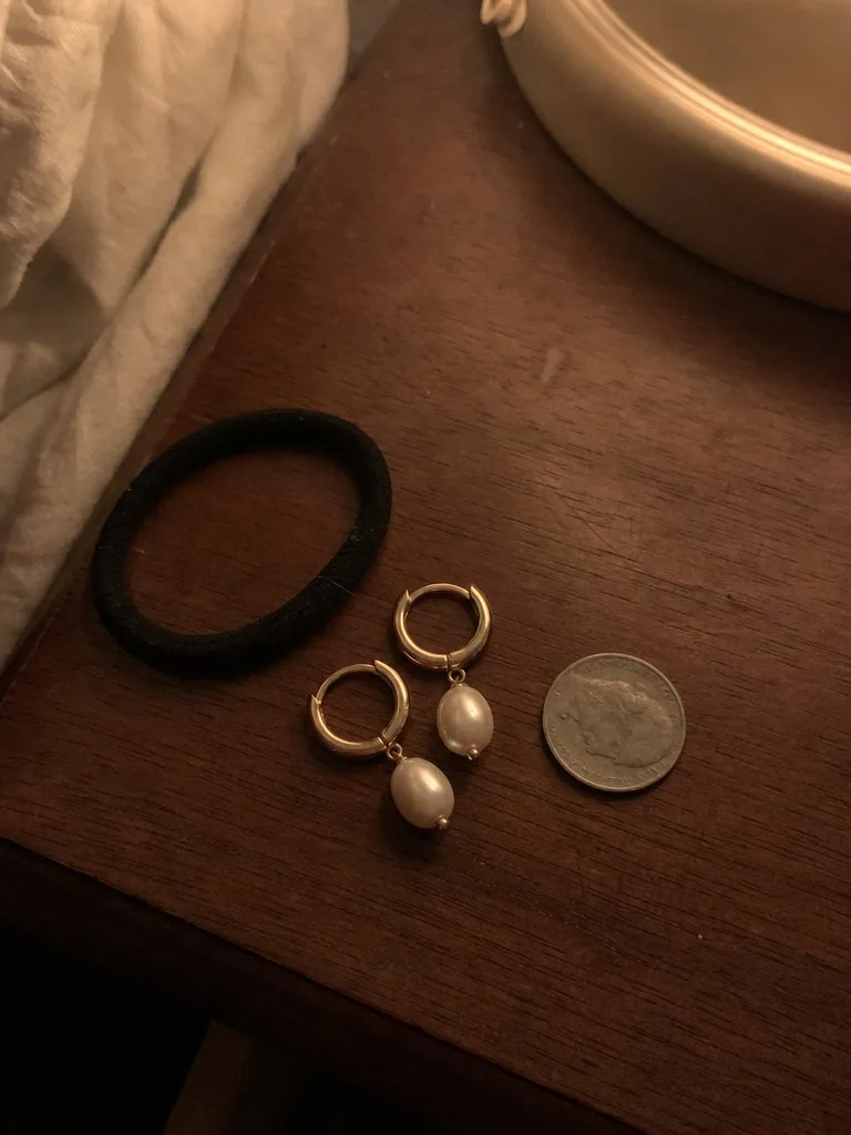 Dim grainy phone photo of a pair of small gold hoop earrings with pearl drops lying on a wooden bedside table next to a hair tie and a coin