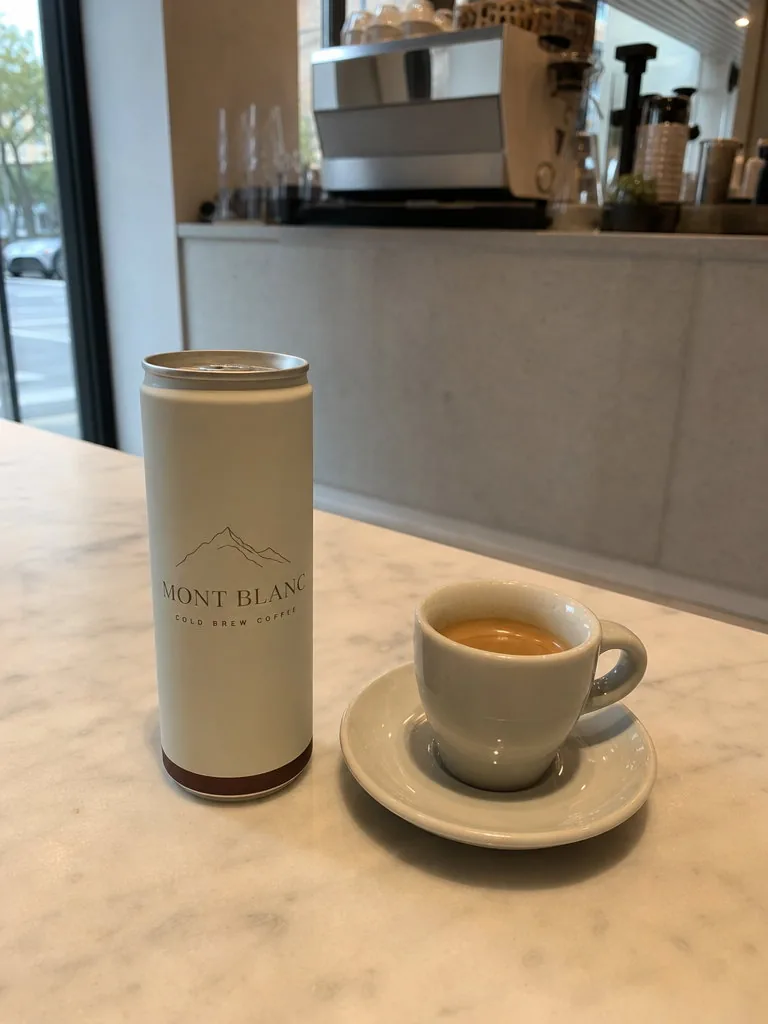 Phone photo of a slim matte cream can printed “Mont Blanc · Cold Brew Coffee” standing on a white marble café table beside an espresso cup, soft window light, the café counter blurred behind