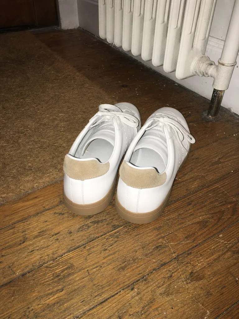 Flash phone photo of a pair of white leather sneakers with gum soles and tan suede heel tabs on a scuffed wooden hallway floor beside a doormat and a radiator