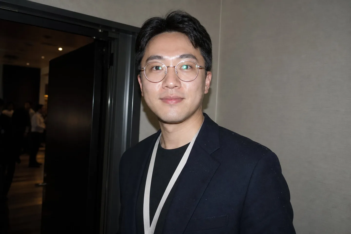 Flash snapshot of a young man with rimmed glasses and a lanyard, white glare across both lenses, shine on his forehead, and lint on a dark blazer
