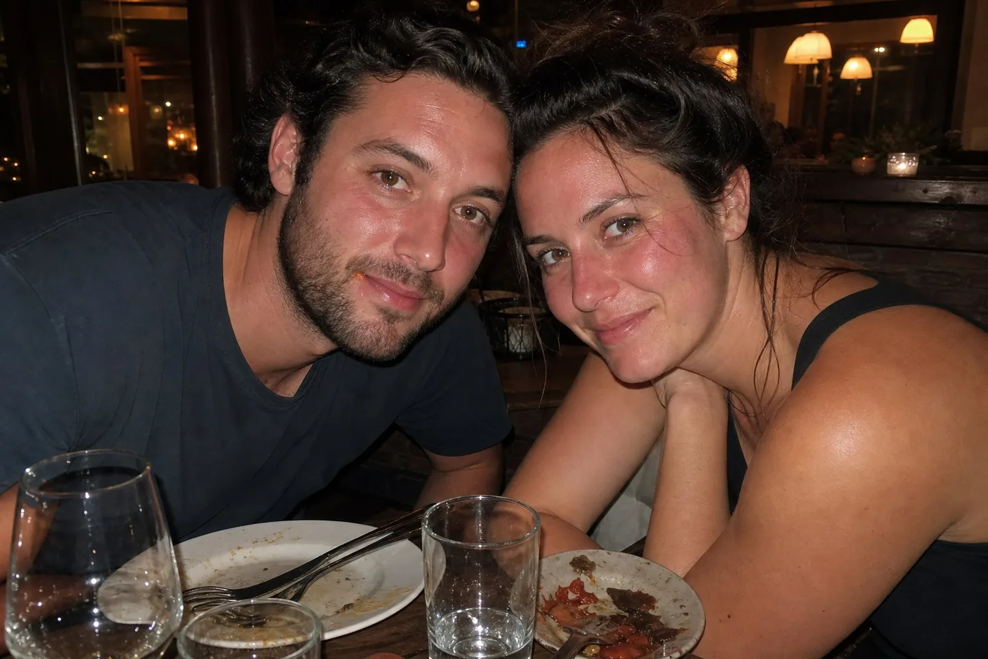 Flash photo of two friends in their thirties leaning in at a restaurant table at night, with hot shine on their faces, red flushed cheeks, stray hairs, and a sauce smear by the man's lip