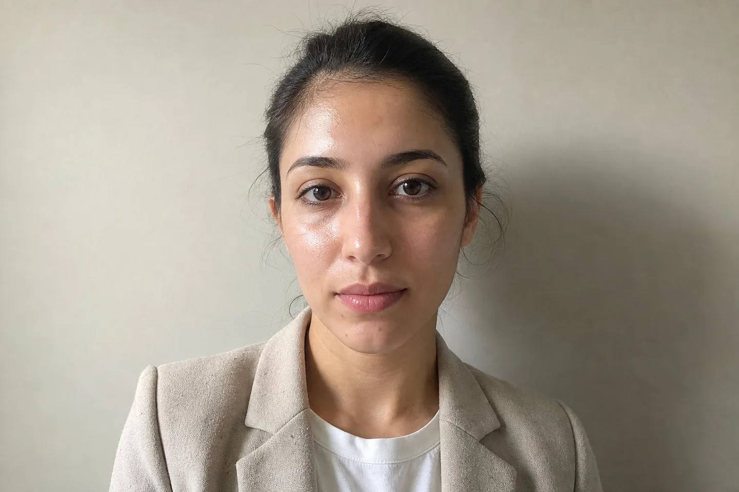 Phone photo of a woman in her late twenties in a light blazer against a plain office wall, with shine on her forehead, a couple of blemishes, dark circles, and lint on the lapel