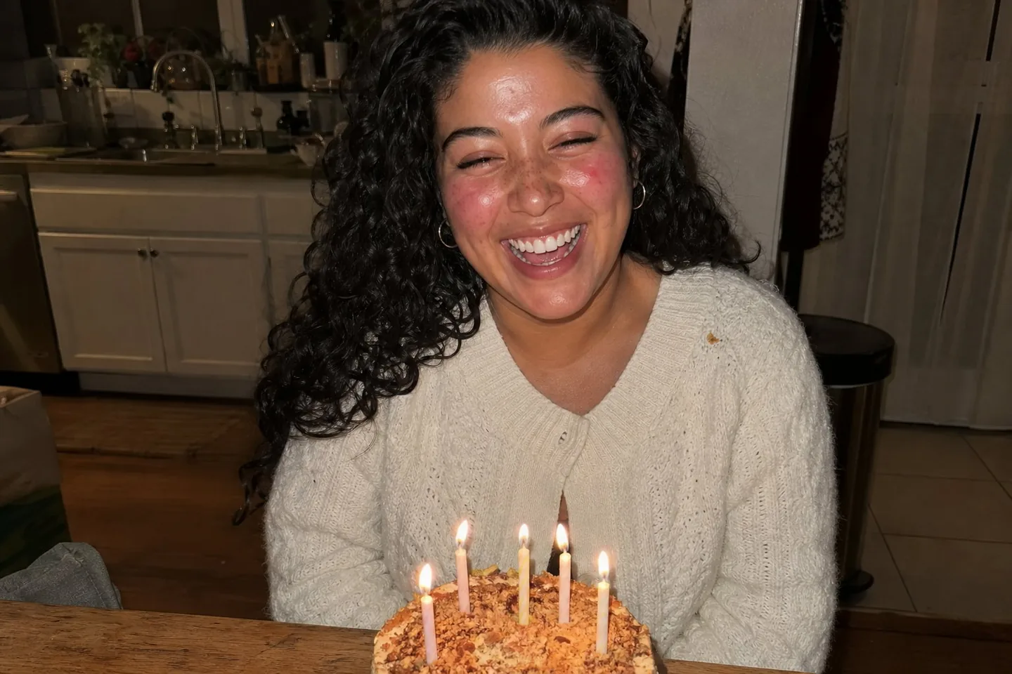 Flash snapshot of a laughing young woman with dark curls and freckles at a kitchen table with a birthday cake, with blotchy red cheeks, shine on her forehead, and a crumb on her cardigan