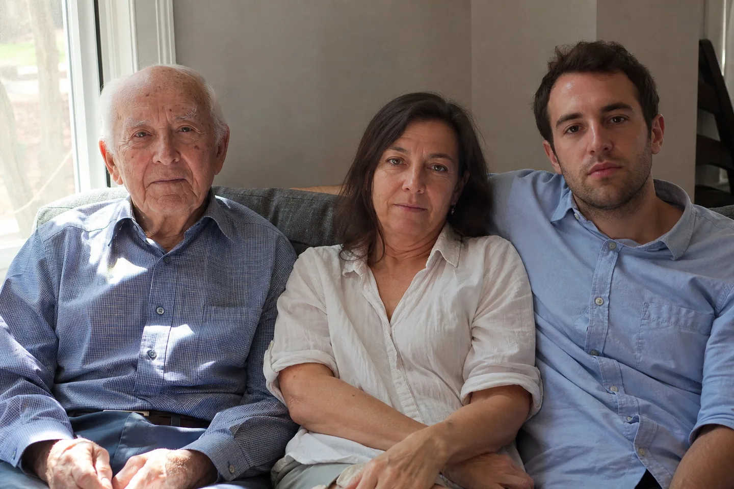 Phone photo of a grandfather, a mother, and her adult son on a sofa by a window, with uneven light across the faces, shine on noses and foreheads, tired eyes, and creased shirts