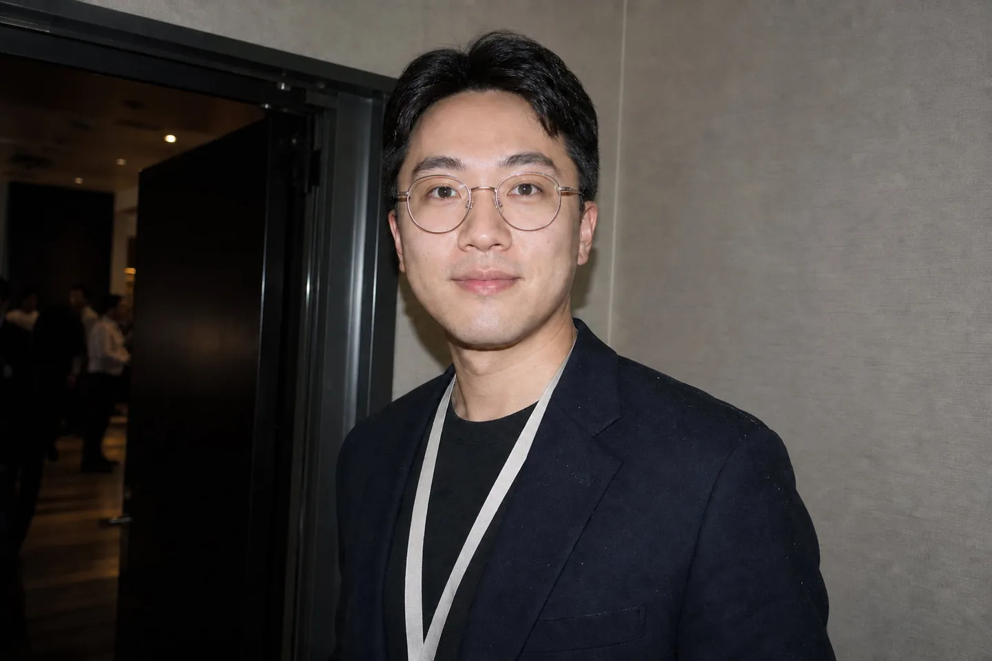 The same conference photo with the lens glare reduced so his eyes read clearly, the flash shine on his forehead toned down, and the lint gone from the blazer