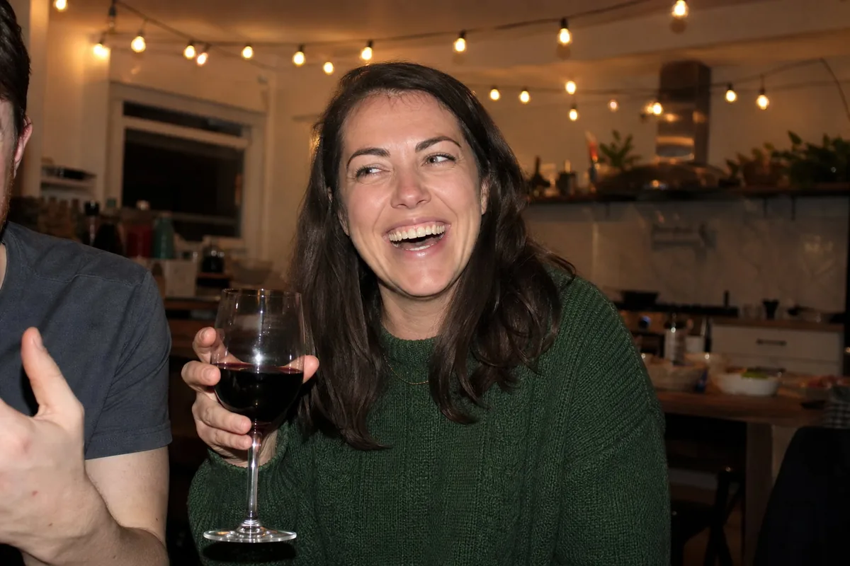 Flash snapshot at a dinner party of a woman in a green knit sweater laughing with a glass of wine, another guest’s shoulder and hand cropped at the frame edge