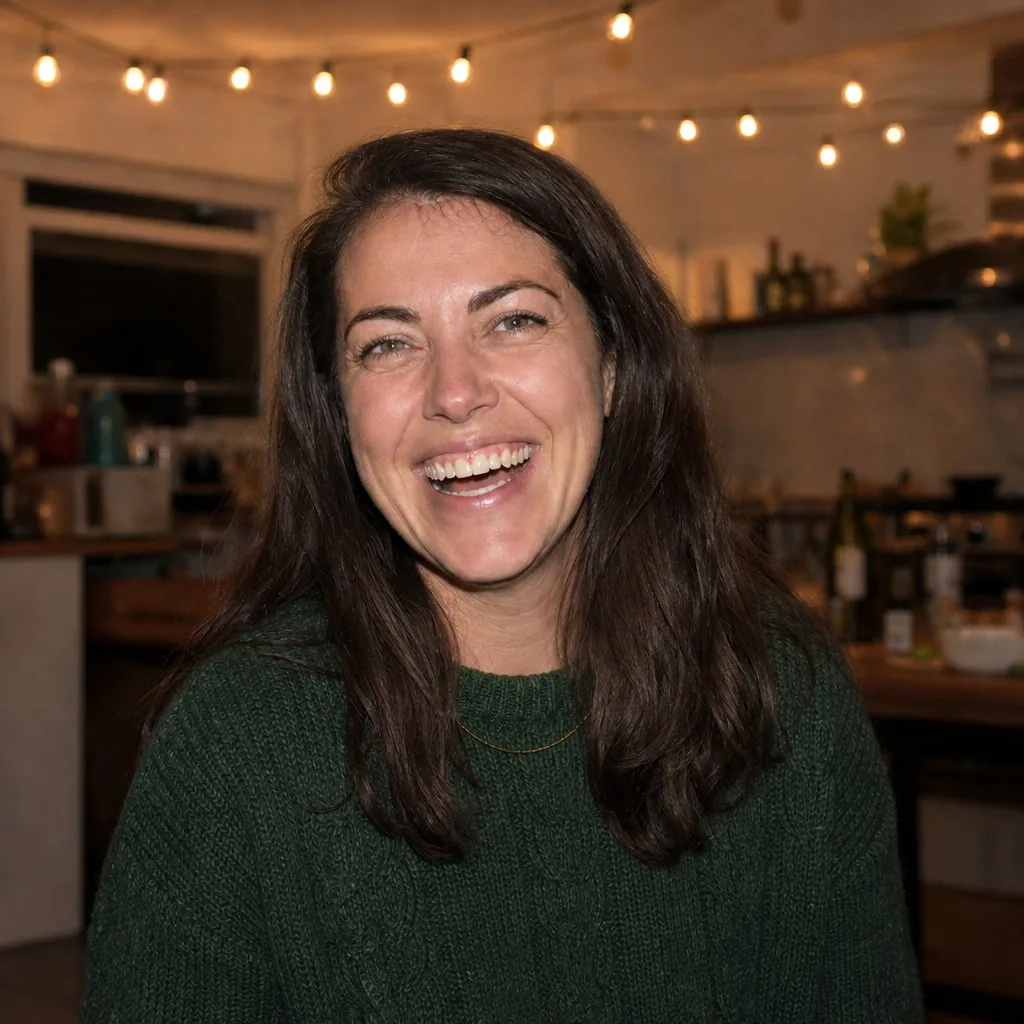 Square profile picture of the same woman laughing in the green sweater, the wine glass and the other guests gone, string lights and kitchen behind her