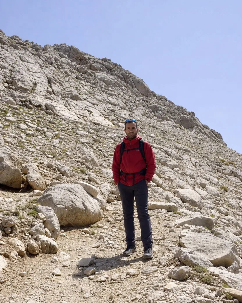 Full-body travel photo of a man in a red windbreaker and backpack standing small on a rocky mountain trail under a harsh midday sun