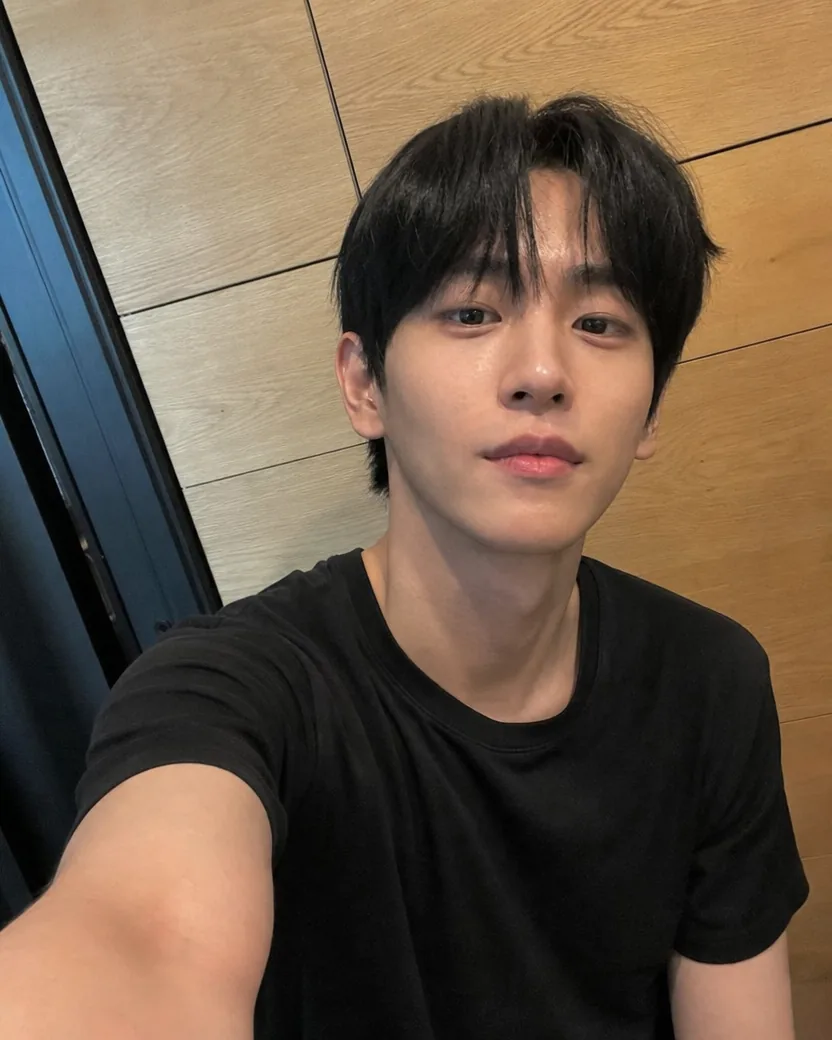 Tilted arm’s-length phone selfie of a young man with a black curtain-fringe haircut in a black T-shirt against an oak-panelled wall, the panels and the door frame leaning with the phone