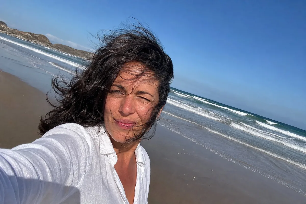 Arm's-length selfie of a woman on a windy beach with dark wavy hair blown across her face, squinting at harsh noon sun in a white linen shirt, tilted horizon, surf and wet sand behind